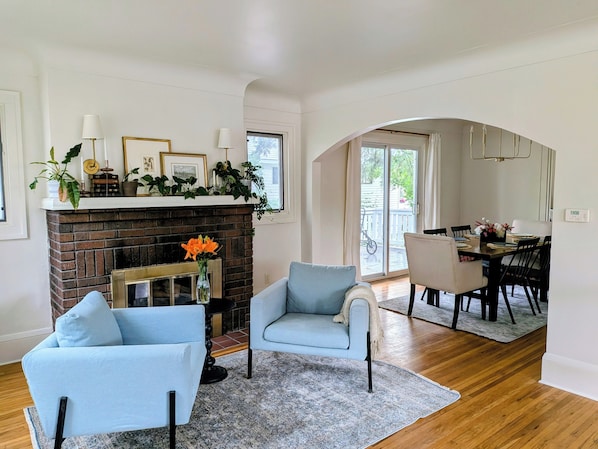A living room with a fireplace, two armchairs, a dining area with a table and chairs, and a view through an archway to the outside.
