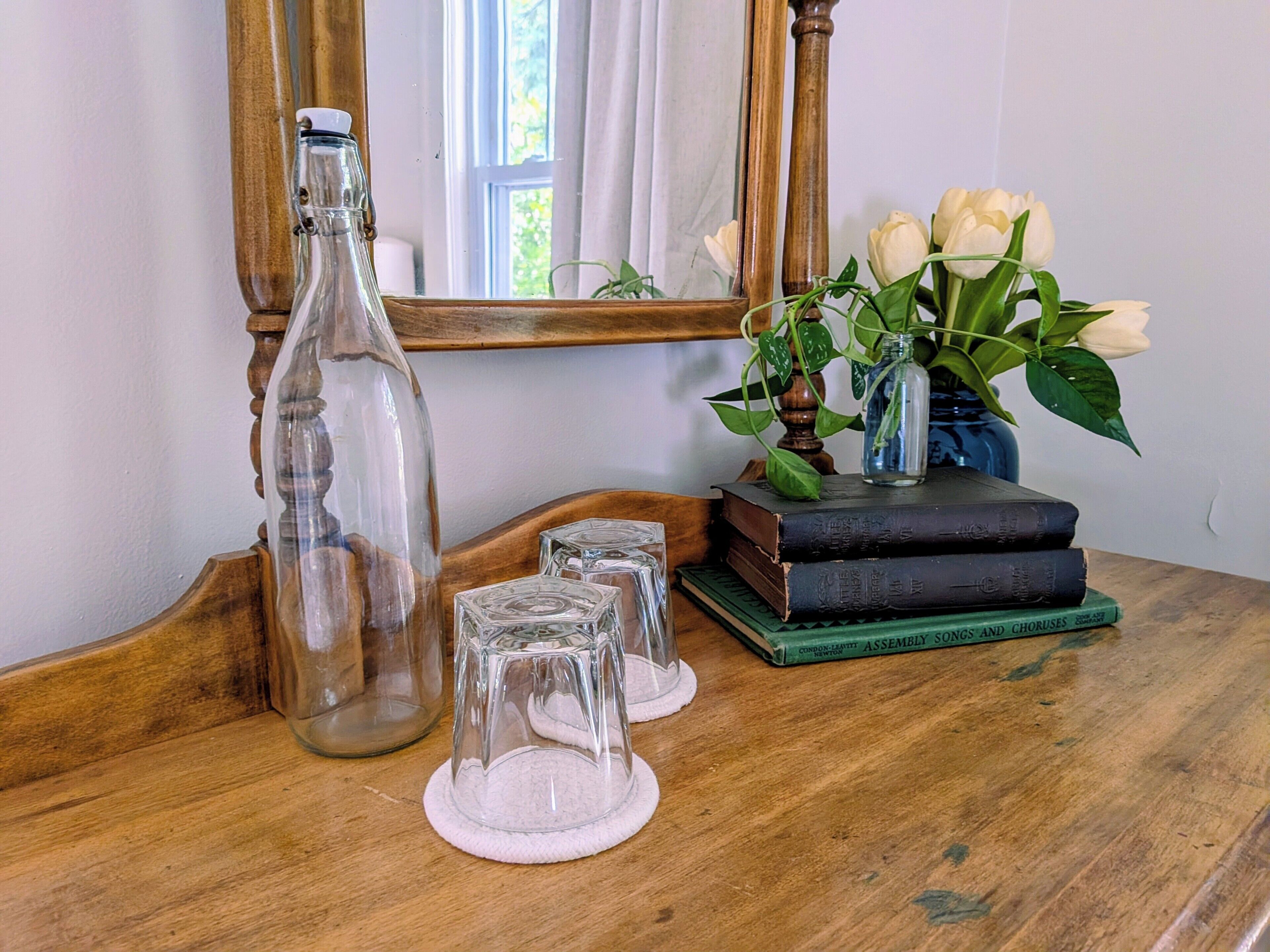 Une table en bois, une bouteille en verre, deux verres transparents, un vase avec des fleurs blanches et une pile de livres.