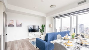 Ein modernes Wohnzimmer mit einer Couch, einem Esstisch für vier Personen, einem Flachbildfernseher und einem großen Fenster mit Blick auf die Stadt.