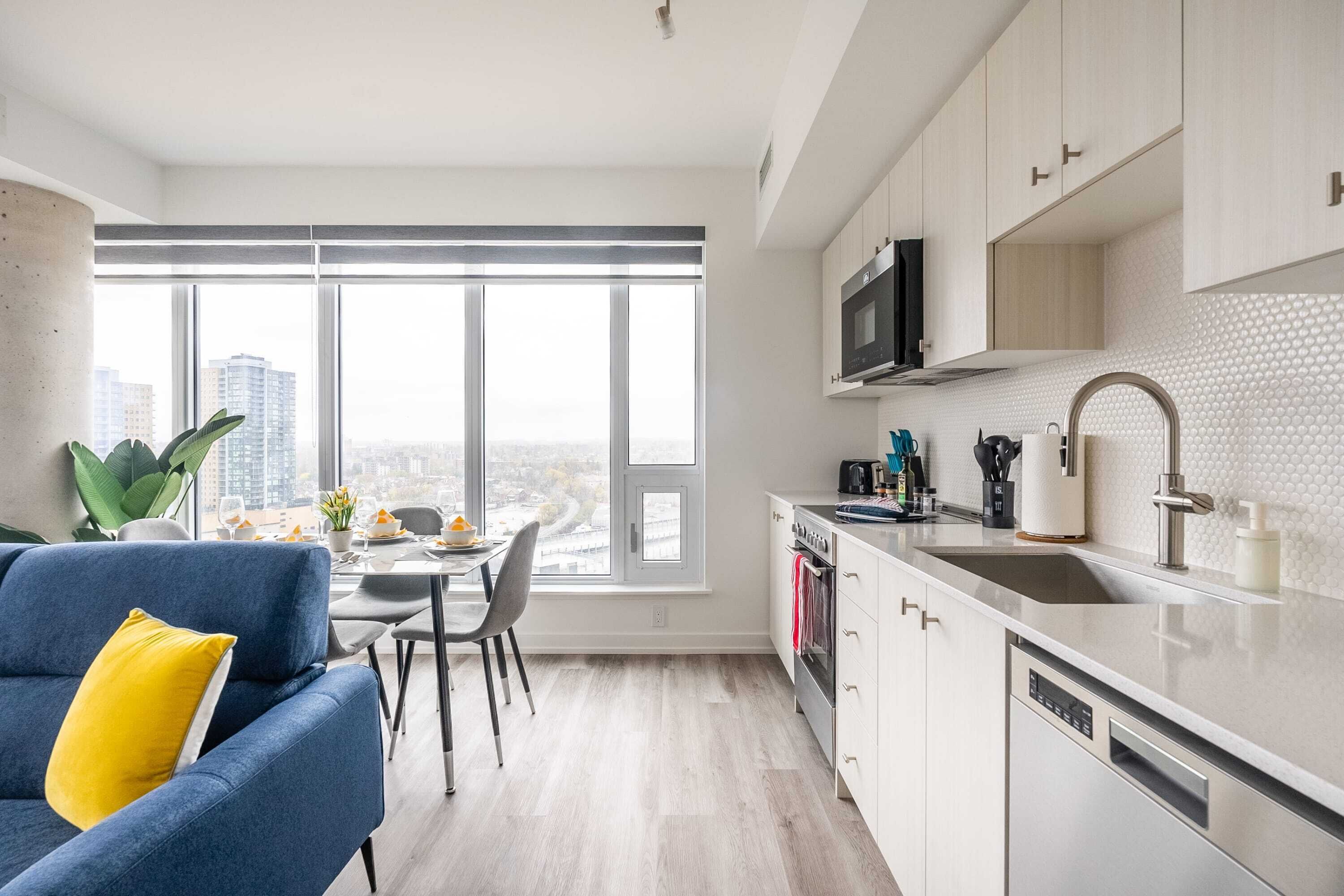 Une cuisine moderne avec une grande fenêtre, des meubles blancs et un canapé bleu agrémenté d’un coussin jaune.