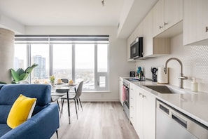 A modern kitchen with a large window, white cabinets, and a blue sofa with a yellow pillow. - High Rise 1BR Condo w Balcony & Beautiful Views (Kitchener)