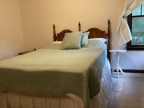 A neatly made bed with a quilted bedspread and pillows, a bedside table with a lamp, and a window with curtains. - Wooded and secluded weekend home; lake is 5 minutes down the hill. (White Bear Lake)