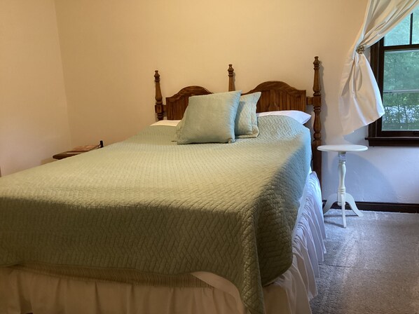 A neatly made bed with a quilted bedspread and pillows, a bedside table with a lamp, and a window with curtains.