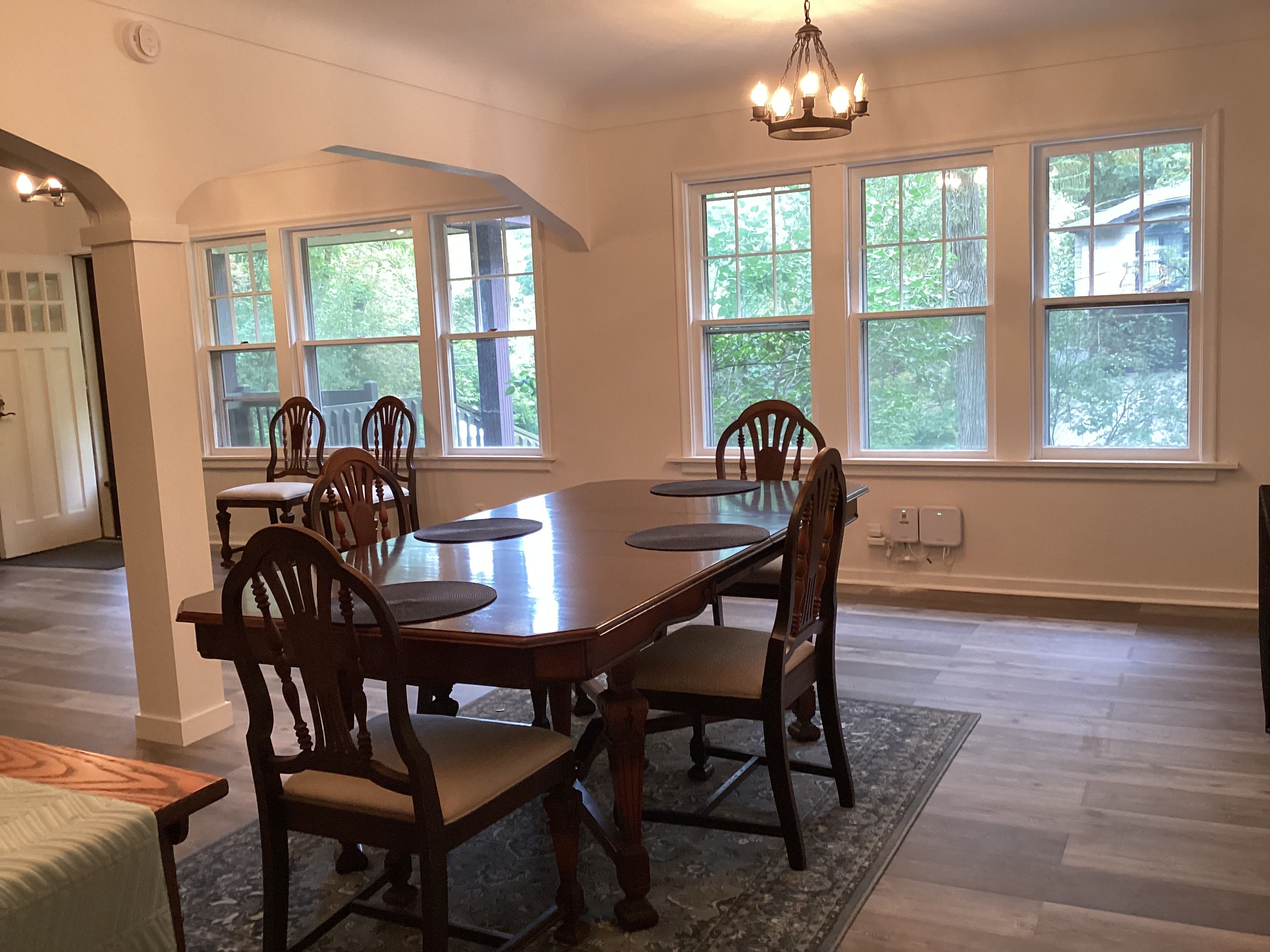 Une salle à manger avec une grande table en bois et six chaises, de grandes fenêtres donnant sur un paysage verdoyant et un lustre.