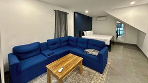 Una habitación moderna con un sofá seccional, una mesa de centro de madera, una cama con ropa de cama blanca y vistas al exterior por una ventana.
