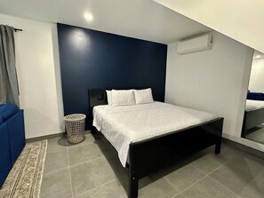 Un dormitorio con cama, pared azul, piso gris y pared blanca, además de una ventana.