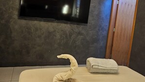 Ein modernes Schlafzimmer mit flachbildfernseher an der Wand, einem ordentlich bezogenen Bett mit Handtüchern und einer Holztür.