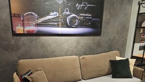 Ein beiges Sofa mit einer Decke und Kissen, ein Flachbildfernseher an einer strukturierten Wand und ein Fliesenboden.