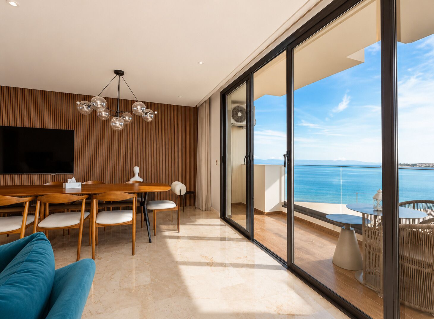 A modern living room with a wooden wall panel, a flat-screen TV, a dining table with chairs, a sofa, and a balcony with a view of the sea.