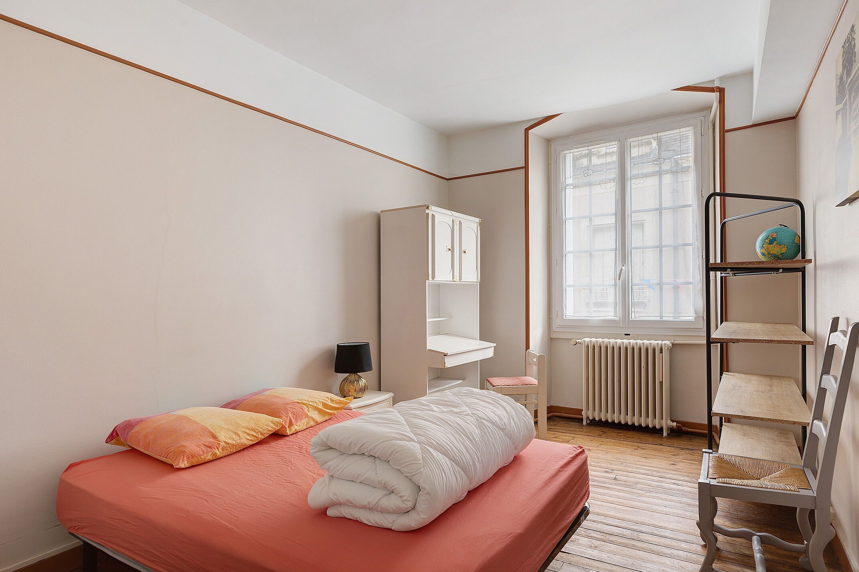 A bedroom with a bed, a nightstand, a lamp, a wardrobe, a window with a view, and a shelf with a globe.