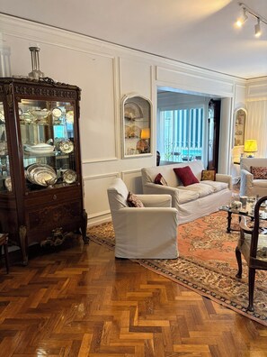 A room with a herringbone parquet floor, a white sofa with patterned cushions, a wooden cabinet with glass doors, and a view through a window with blue and white striped curtains. - Spacious Recoleta apartment in desired location - French style & Modern Comfort (Buenos Aires)