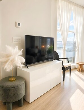 Uma sala de estar moderna com uma televisão de tela plana sobre um móvel branco, um banquinho redondo, uma cadeira preta e vista para o mar através de cortinas leves.