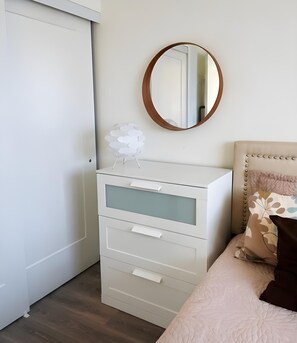 Quarto com cômoda branca, espelho redondo e cama com travesseiro estampado.