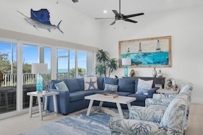 A spacious living room with a large blue sofa, a coffee table, and a patterned armchair. There is a ceiling fan, a map painting, and a view of the ocean through large windows.