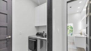 Uma cozinha compacta com forno embutido e micro-ondas, armários brancos e vista para um banheiro por uma porta aberta.