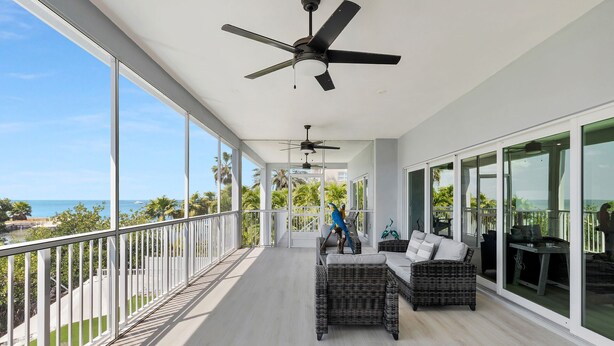 Uma varanda com vista para o oceano, ventiladores de teto, móveis de vime e uma mesa de jantar.