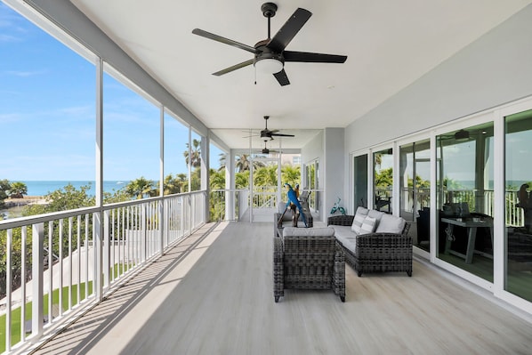 A balcony with a view of the ocean, ceiling fans, wicker furniture, and a dining table.