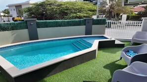 A rectangular pool with clear blue water, surrounded by artificial grass and white chairs. - Caribbean Oasis - Bright - cozy - Close to beaches. 1-bedroom apartment with ac (Noord)