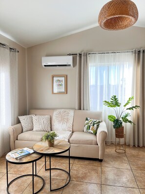 A living room with a beige sofa, two round tables, a potted plant, and a hanging light fixture. - Caribbean Oasis - Bright - cozy - Close to beaches. 1-bedroom apartment with ac (Noord)