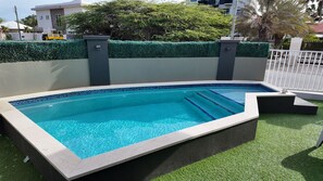Ein rechteckiger Swimmingpool mit einer Fliesenbordsteinkante, umgeben von einer niedrigen Hecke und einem weißen Zaun.