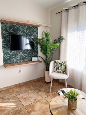 A living room with a flat-screen TV mounted on a wall with a tropical wallpaper, a potted plant, a white armchair with a patterned cushion, and a round coffee table with a plant. - Caribbean Oasis - Bright - cozy - Close to beaches. 1-bedroom apartment with ac (Noord)