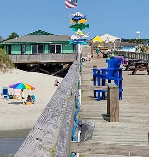 Een houten looppad leidt naar een strand met een redderpost, strandstoelen en een kleurrijke parasol.
