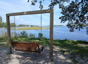 Un columpio de madera con banca, con vistas a un cuerpo de agua bordeado de hierba y árboles.
