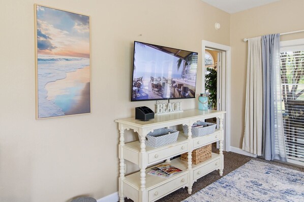 Een kamer met een aan de muur bevestigde televisie, een witte console tafel met opbergmanden en een lijst met een afbeelding van een strand.