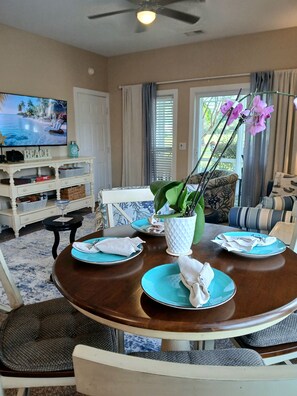 A dining area with a round table set for four, surrounded by chairs. There is a television mounted on the wall, a potted plant on the table, and a decorative item labeled 'BEACH' on the wall. - Prime Sea Trail condo. 1st floor end unit-villa and suite combined.  Remodeled! (Sunset Beach)