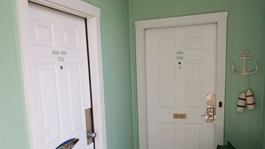 Un pasillo con dos puertas blancas, cada una identificada con un número y nombre de habitación. Hay un felpudo con el texto 'SO NICE TO SEE YOU' y un cartel de bienvenida sobre la puerta.