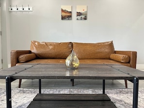 Una sala de estar con un sofá de cuero marrón, una mesa de centro de madera con un jarrón de cristal decorativo y dos cuadros colgados en la pared.
