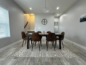 A dining area with a table and chairs, a clock on the wall, and a window with blinds.