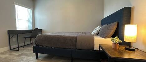A bedroom with a bed, bedside table, lamp, and a desk with chair.