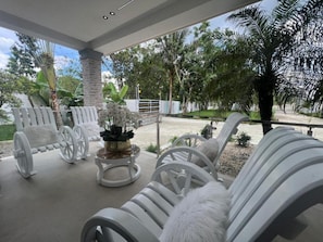 A covered outdoor seating area with white chairs and a central table, surrounded by greenery and palm trees.