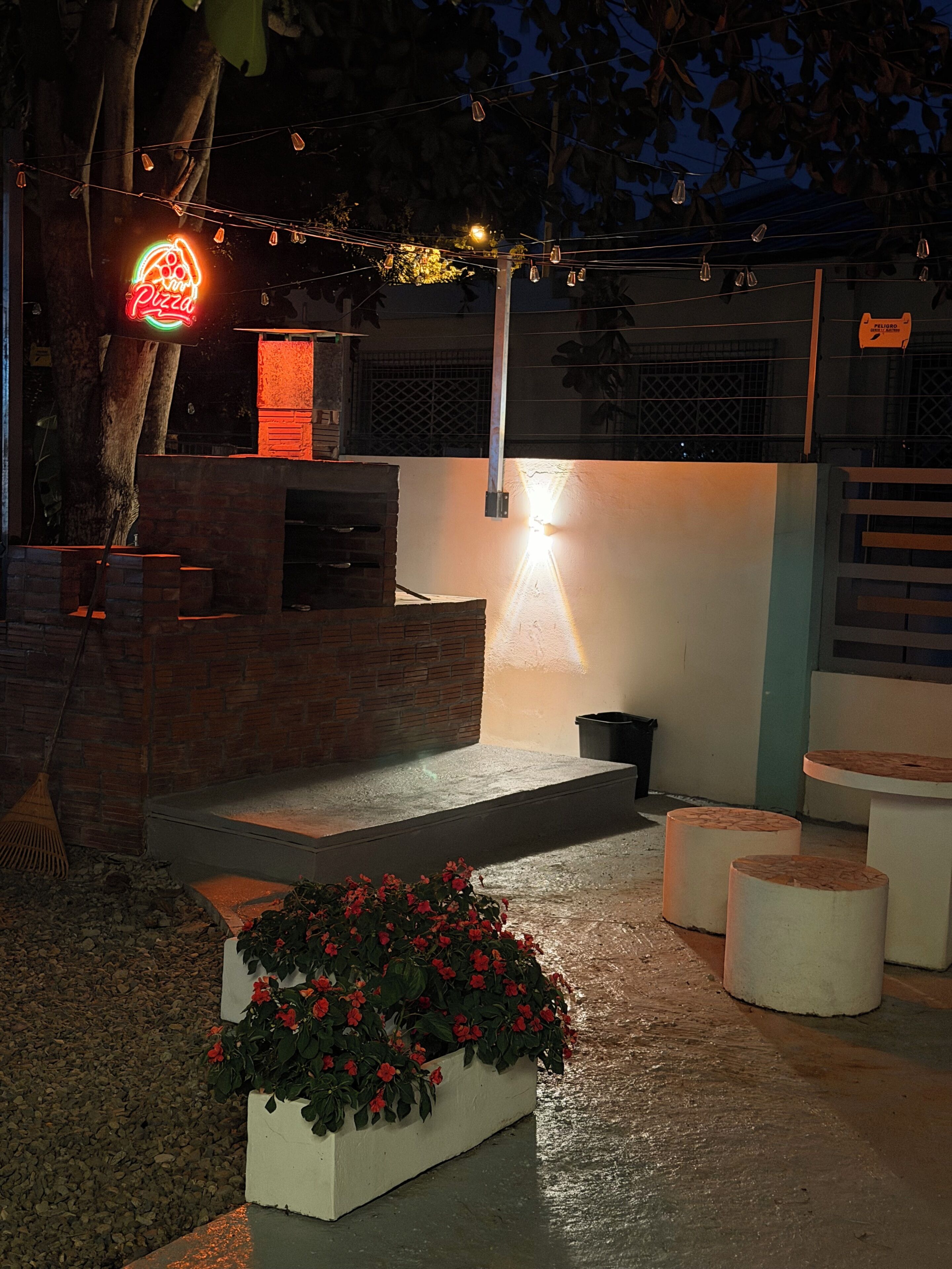 Un patio al aire libre con pared de ladrillo, un letrero de neón con la palabra pizza, una lámpara colgante y una maceta con flores rojas.