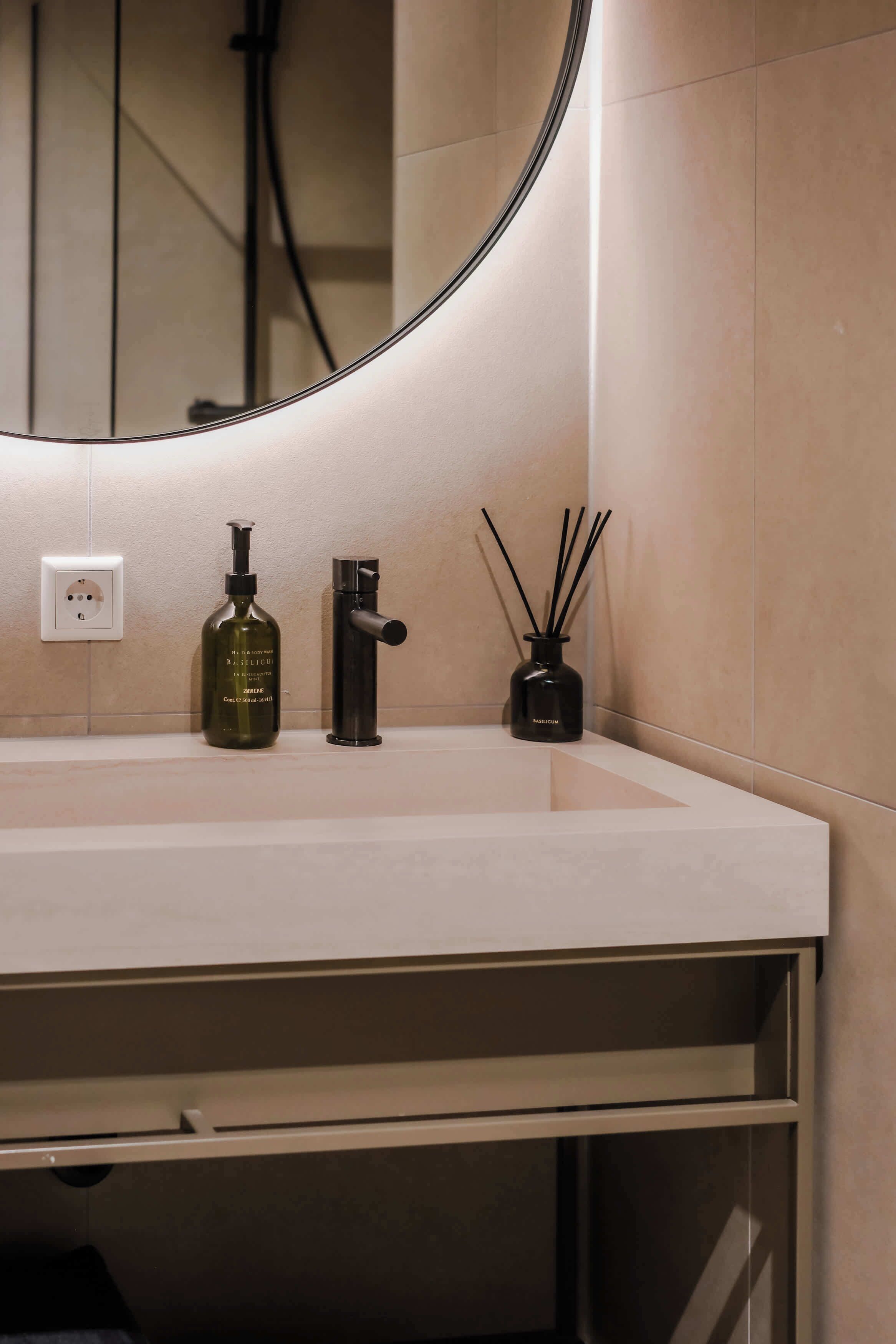 A bathroom with a white sink, a mirror, a bottle of body wash, a faucet, and a reed diffuser.