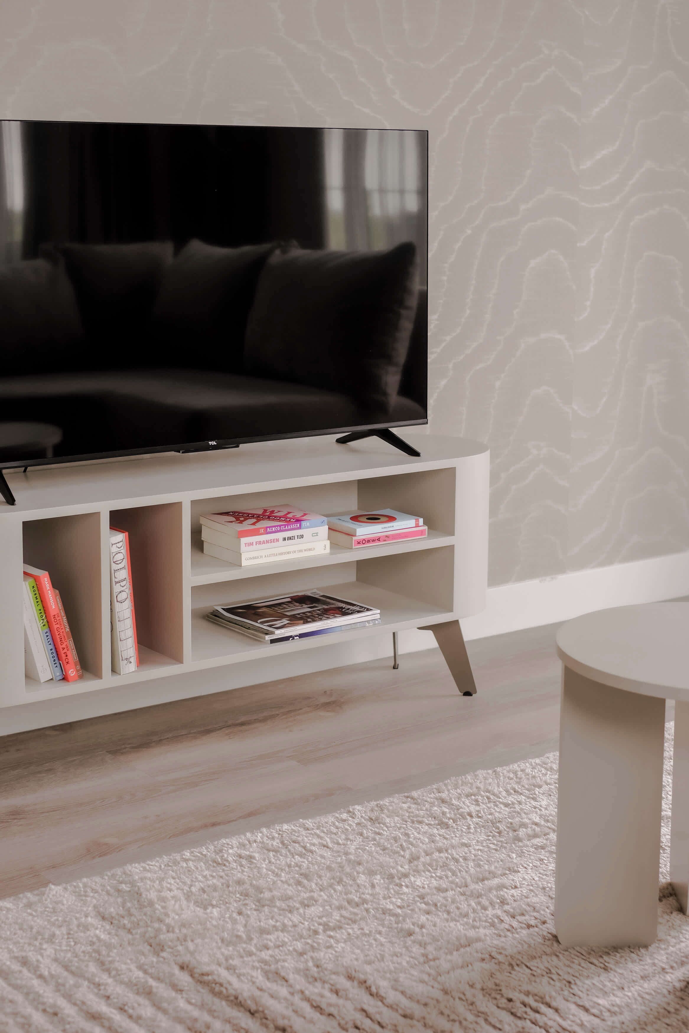 Un salon moderne avec un téléviseur à écran plat posé sur un meuble blanc, un canapé blanc et une table basse blanche. Des livres et des magazines sont disposés sur les étagères situées sous le téléviseur.