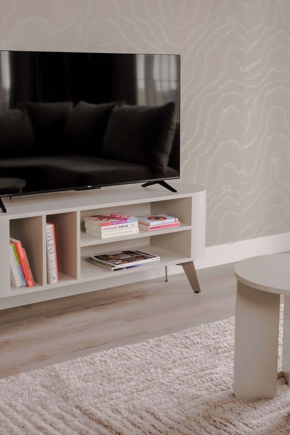 Ein modernes Wohnzimmer mit einem Flachbildfernseher auf einem weißen Schrank, einem weißen Sofa und einem weißen Couchtisch. Unter dem Fernseher befinden sich Regale mit Büchern und Zeitschriften.