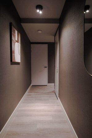 Un pasillo con suelo de madera, una puerta blanca al final y una obra enmarcada en la pared izquierda.