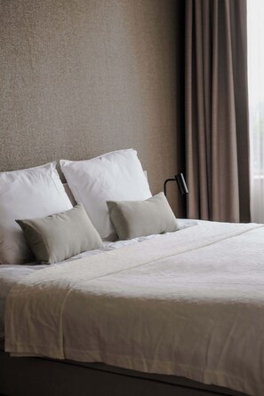 Una cama bien hecha con edredón blanco y varios almohadones, una lámpara de pared negra y una ventana con cortinas.