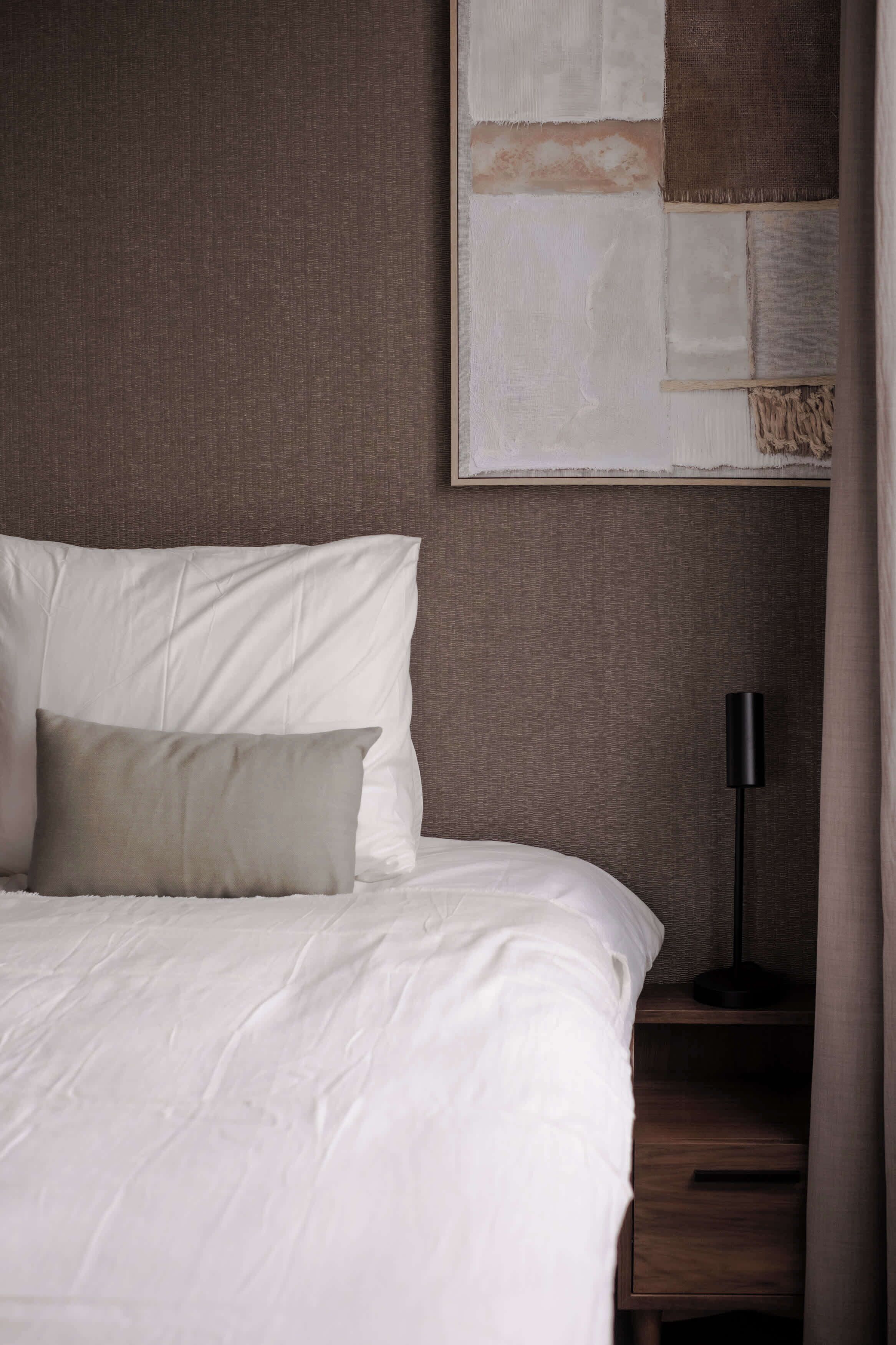 Uma cama bem arrumada com roupa de cama branca e duas almofadas, um abajur de chão escuro e um grande quadro abstrato na parede.