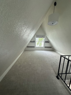 An attic room with a sloped ceiling, a window with a view of greenery, and a black metal railing.