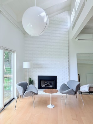 A modern living room with a white brick wall, a fireplace, and two grey armchairs around a round wooden table.