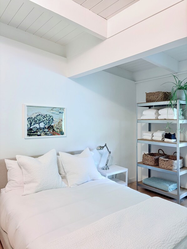 Un dormitorio con cama, mesita de noche, lámpara, estante con toallas y cestas, y un cuadro colgado en la pared.