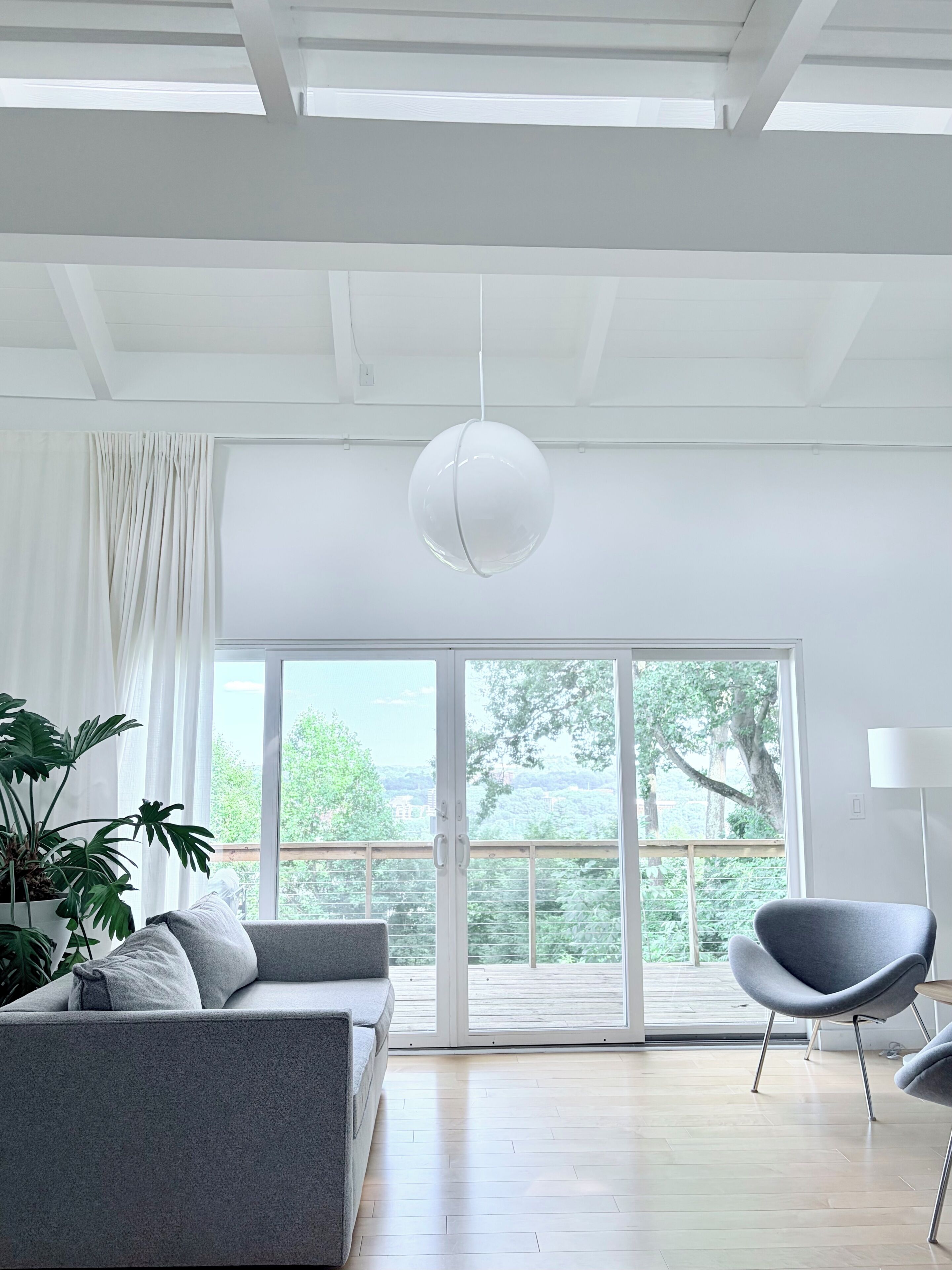 A modern living room with a grey sofa, a grey armchair, a hanging light fixture, and a large window with a view of greenery and buildings.