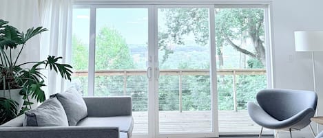 A modern living room with a grey sofa, a grey armchair, a hanging light fixture, and a large window with a view of greenery and buildings.