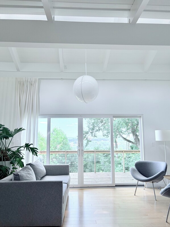 Un salon moderne avec un canapé gris, un fauteuil gris, un luminaire suspendu et une grande fenêtre donnant sur un paysage verdoyant et des bâtiments.