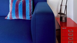 Ein Zimmer mit einem blauen Sofa, einem gestreiften Kissen, einem gerahmten abstrakten Gemälde, einer schwarzen Schreibtischlampe und einem roten Stauraum mit Schubladen.