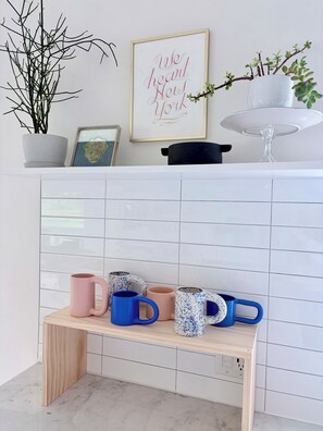 Un área de cocina con salpicadero de azulejos, un banco de madera con tazas, una planta en maceta y un letrero enmarcado.