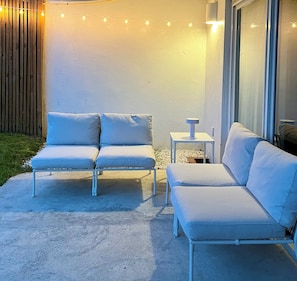 Un espacio al aire libre con dos sofás blancos acolchados y una pequeña mesa, frente a una pared blanca decorada con luces de cuerda.